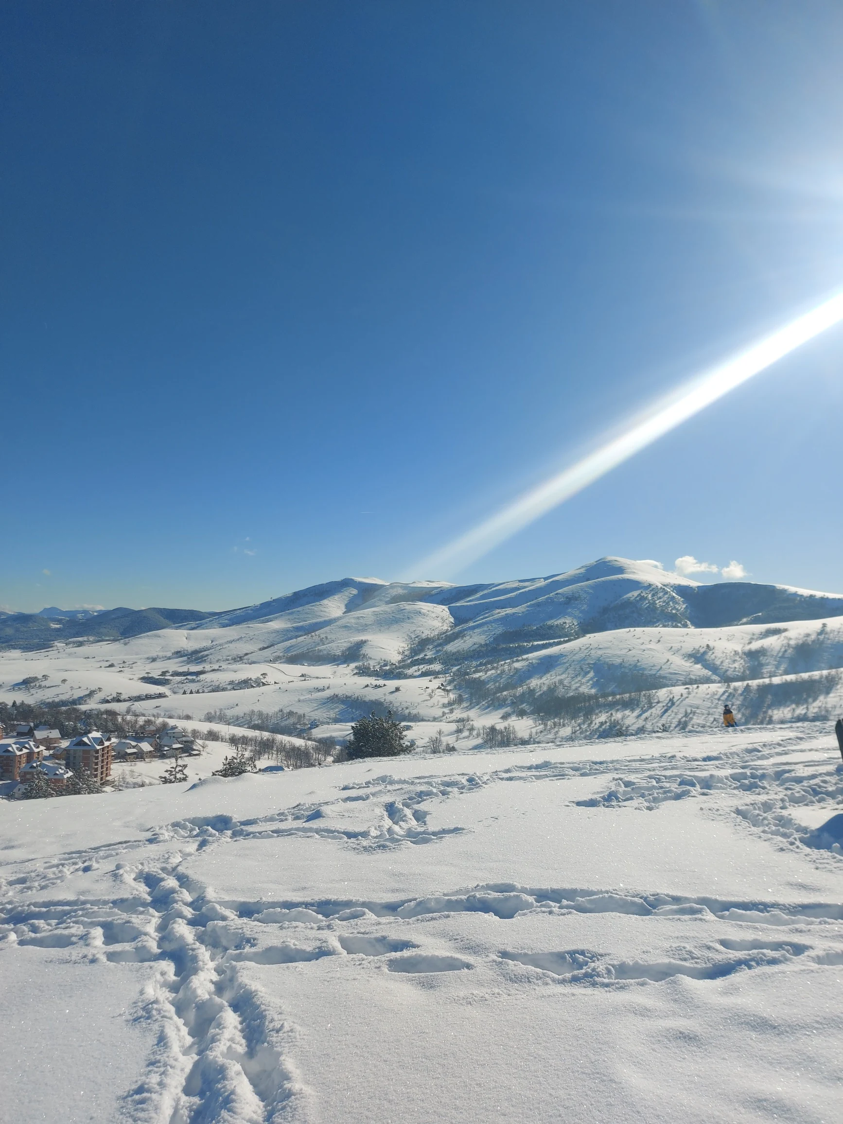 Wide snow-covered landscape with mountain peaks in the background, visible footprints in the snow, and a clear blue sky with sun rays. Distant buildings are visible on the left side.