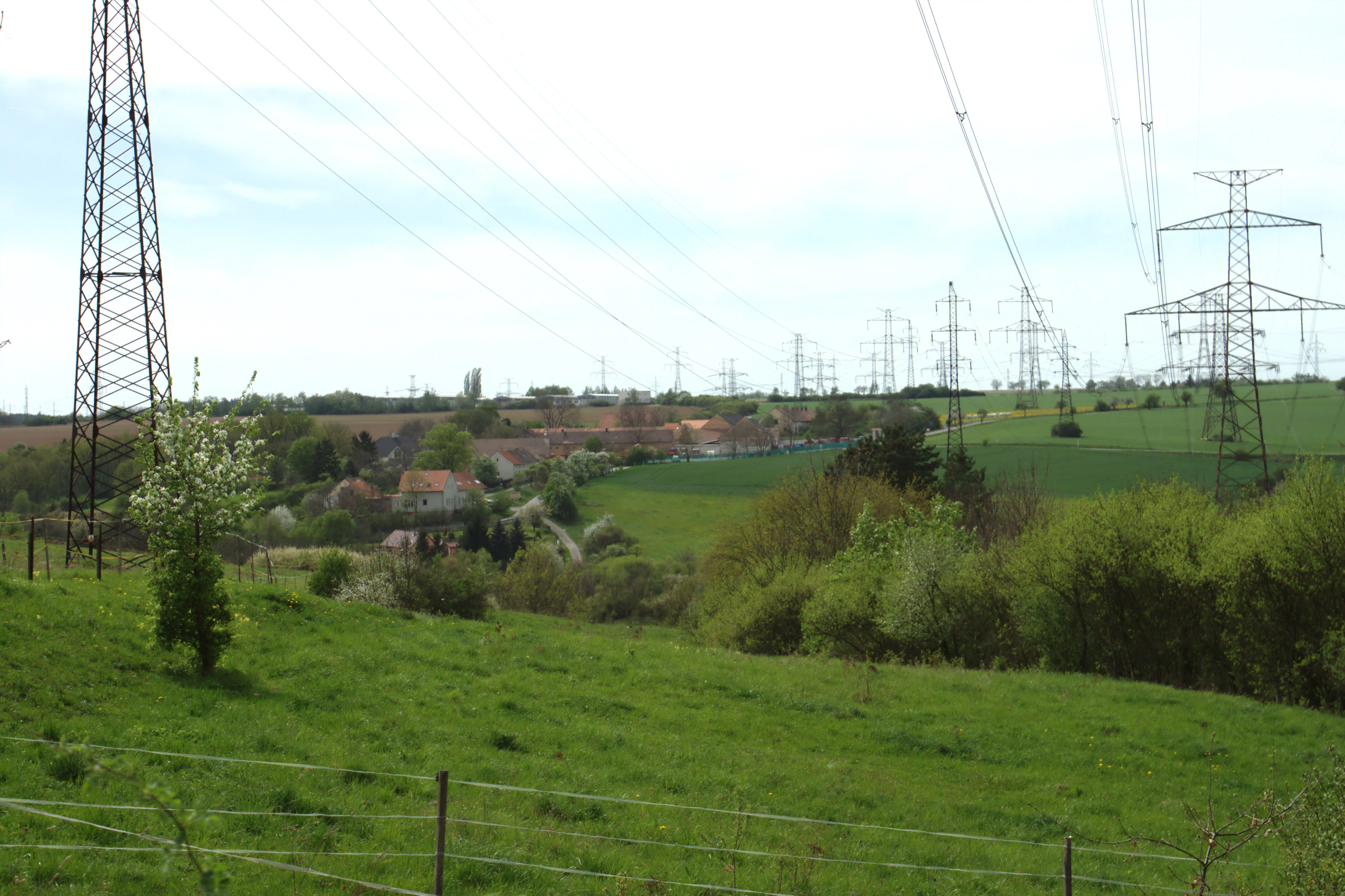 A wide view of green meadows and rolling hills with several large metal electrical transmission towers in the foreground and distance, near a rural settlement.