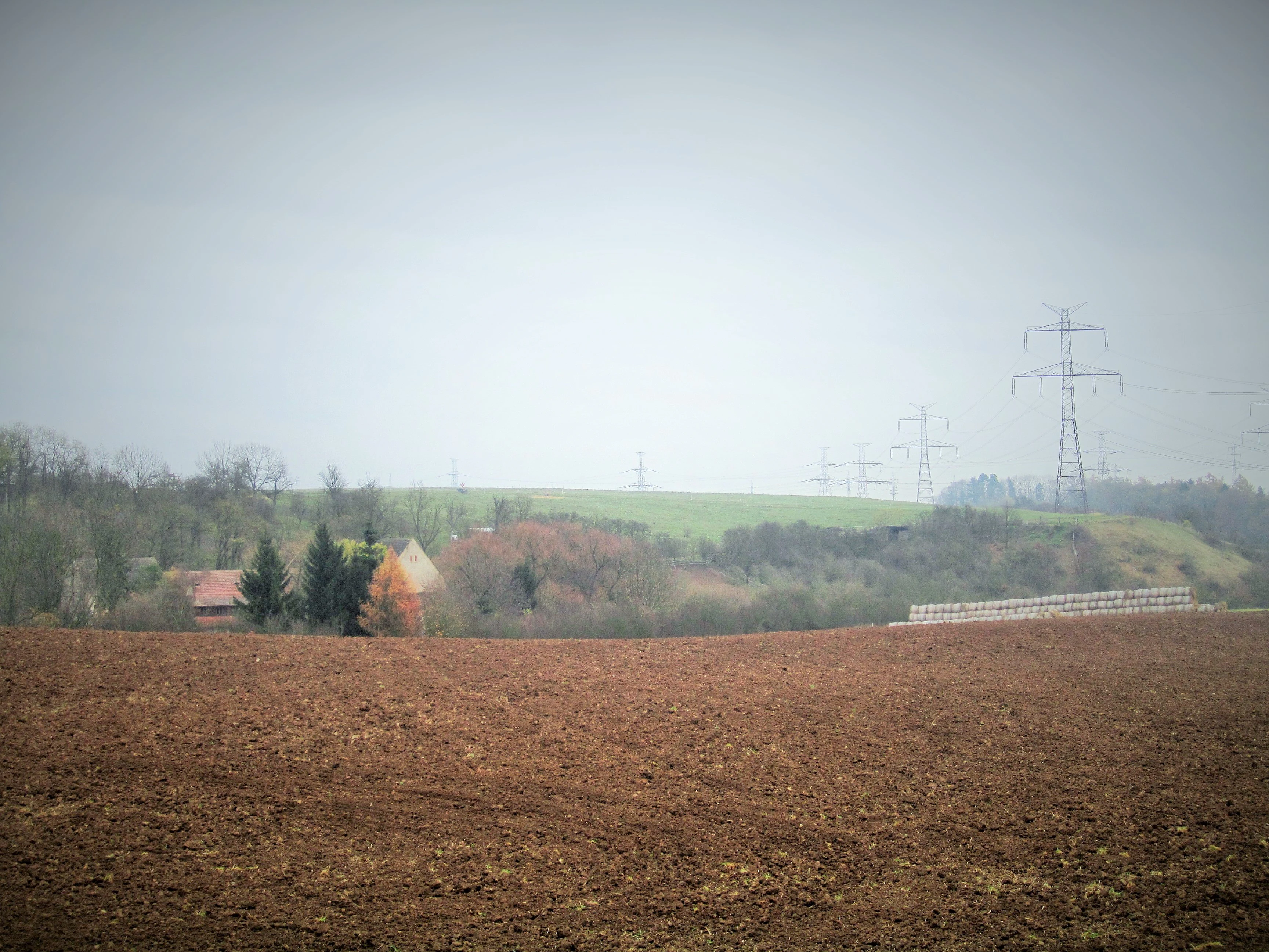 A wide landscape view of an agricultural field with brown, plowed earth in the foreground and a distant valley with trees, structures, and power lines under a hazy, overcast sky.
