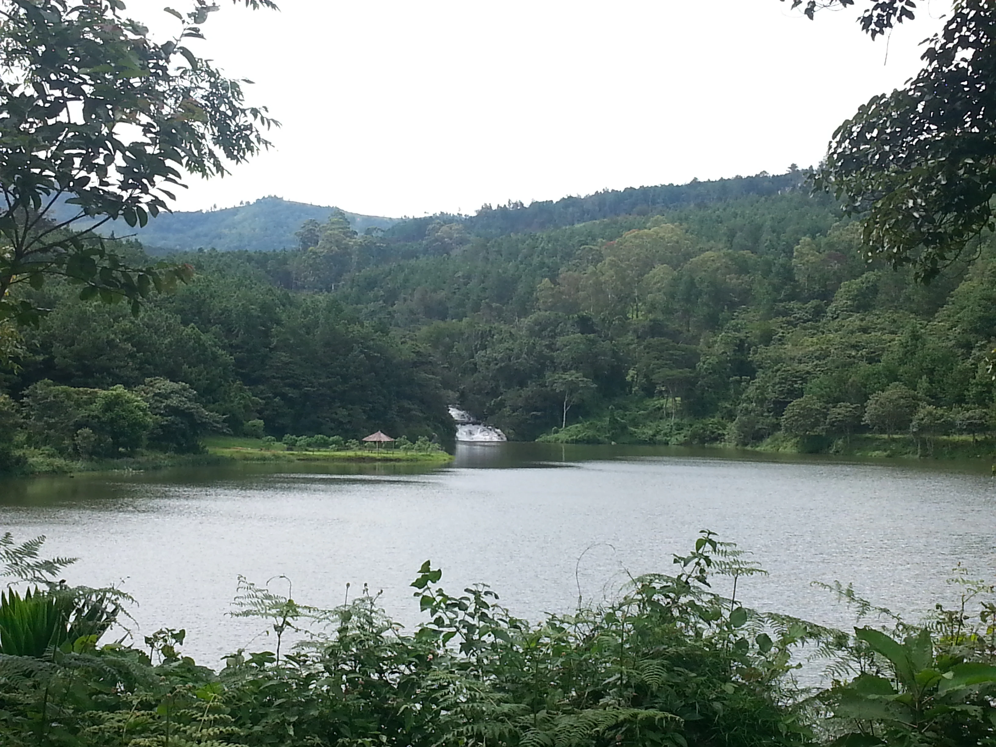 A wide shot of a calm reservoir lake surrounded by dense, lush forest and mountains, with a small waterfall visible in the distance.