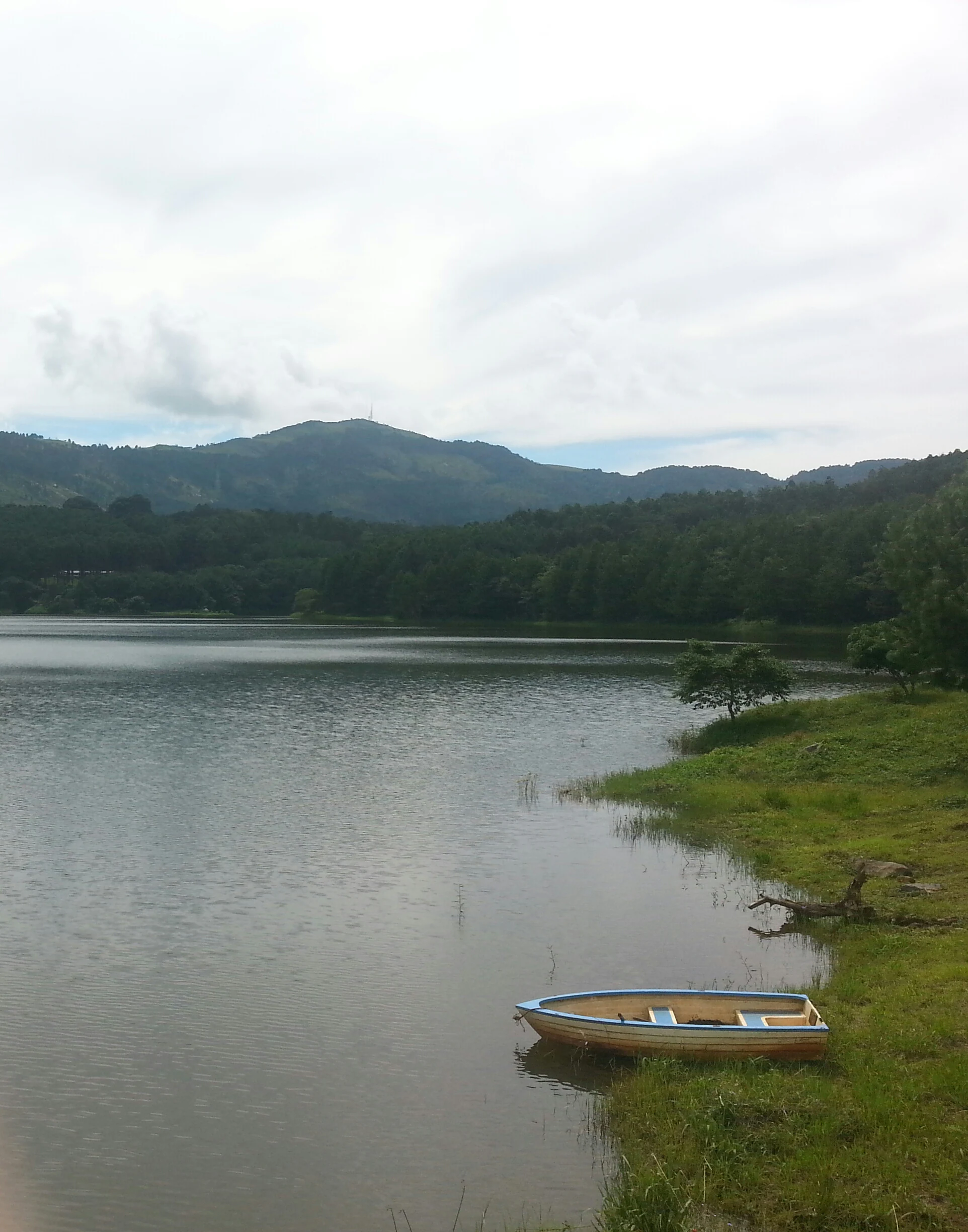 A small, light-colored boat rests in the calm water at the grassy edge of a reservoir with forested hills in the background.