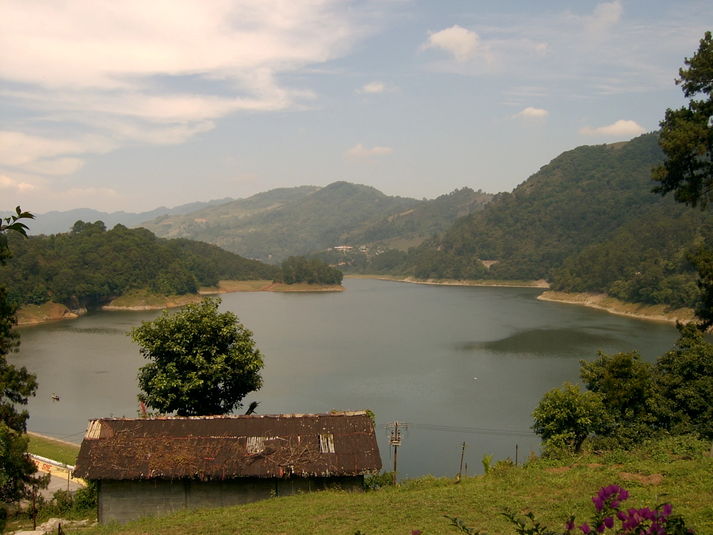 A wide shot showing a large reservoir created by the Necaxa River, surrounded by rolling, forest-covered mountains under a partly cloudy sky. A small, simple structure with a dark roof sits in the foreground.