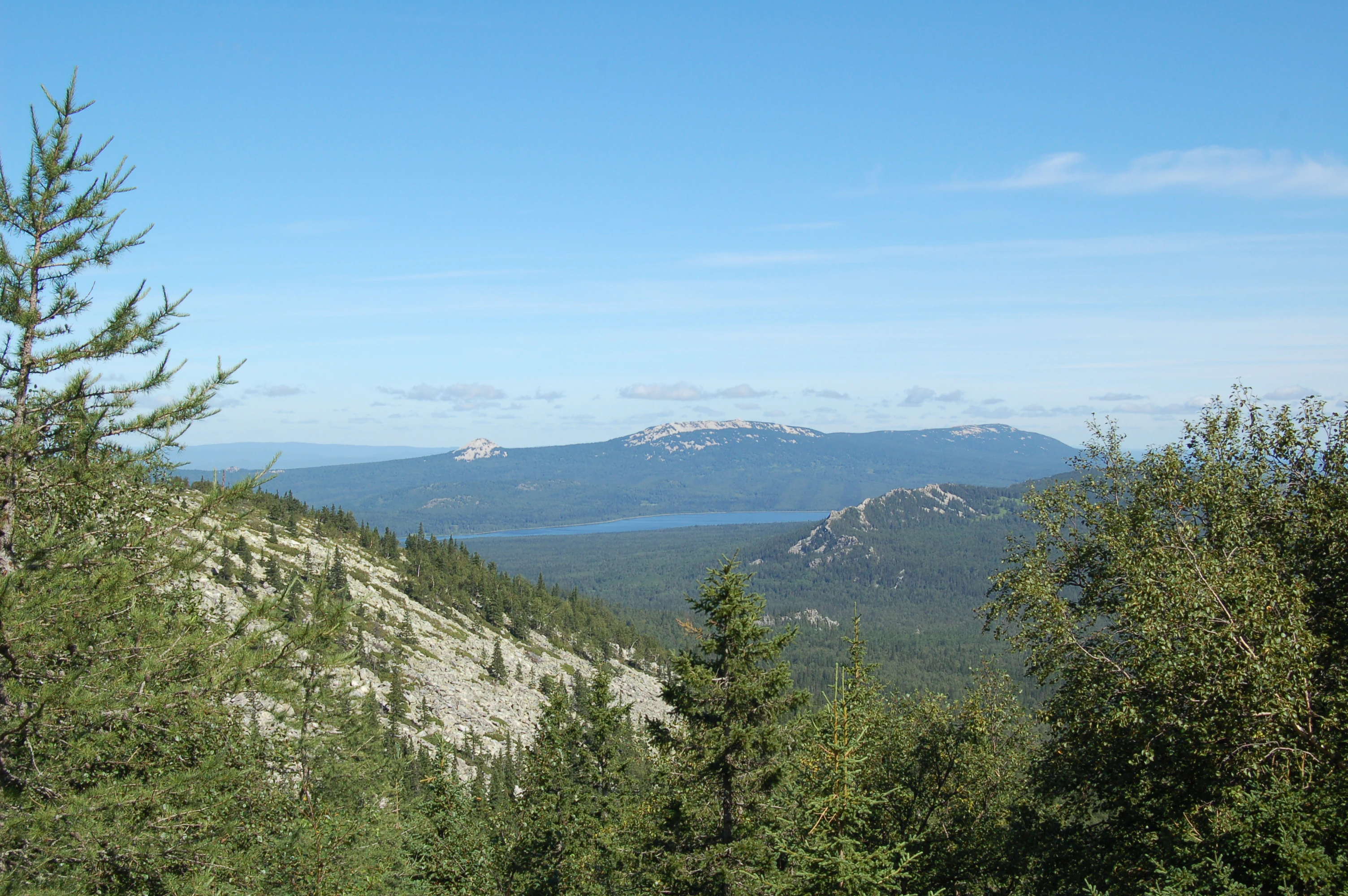 A wide mountain vista overlooking a lake nestled between dense evergreen forests and mountain ridges under a clear blue sky.
