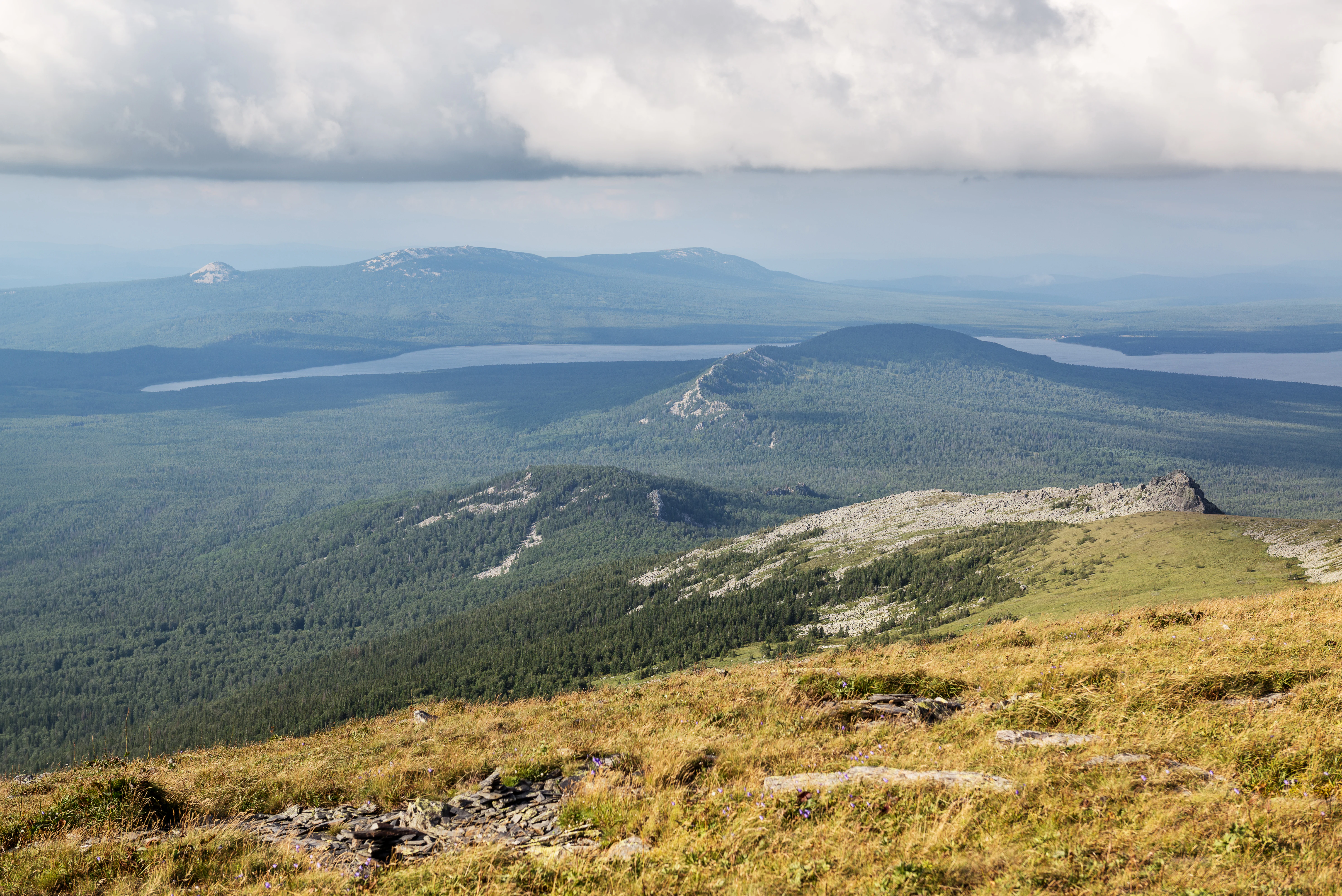 Panoramic landscape view of Zyuratkul National Park with grassy hills, forests, mountain ranges, and a lake in the distance under a cloudy sky